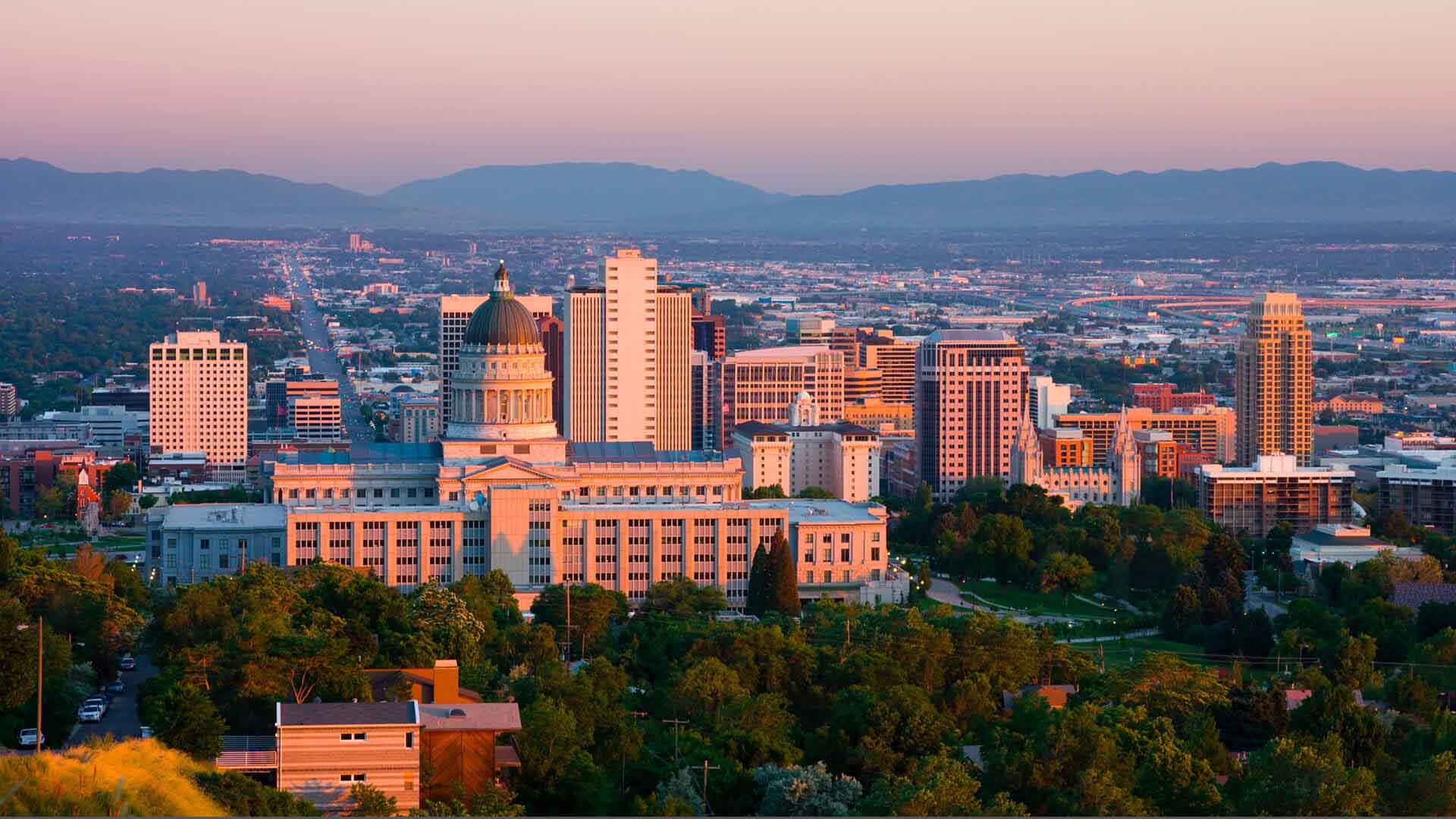 Utah State Capitol overlooks Salt Lake City buildings and trees beneath distant mountains at sunset.